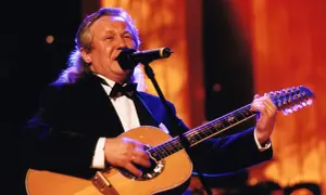 Artysta w czarnym smokingu gra na gitarze, śpiewając do mikrofonu. W tle pomarańczowe światła, jakby na scenie, gdzie kiedyś występowały siedem dziewcząt z Albatrosa.