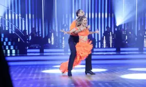 Tancerze w pomarańczowych strojach wykonują dynamiczny big dance. W tle orkiestra, a na scenie migoczą niebieskie światła, jakby sypały się pieniążki.