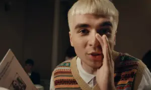 Młody mężczyzna z blond włosami, w kamizelce w kolorowe wzory, trzyma w ręku kartkę z tekstem, jakby czytał papuga.