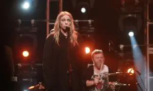 Wokalistka z blond włosami śpiewa do mikrofonu, a za nią perkusista gra na bębnach. To jak hey teksański tekst na scenie.
