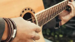 Dłoń grająca na gitarze, tworząc akord C. Widać bransoletę na nadgarstku i fragment pudła rezonansowego.