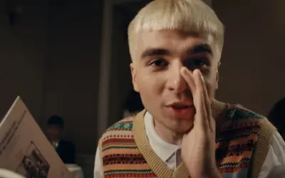 Młody mężczyzna z blond włosami, w kamizelce w kolorowe wzory, trzyma w ręku kartkę z tekstem, jakby czytał papuga.