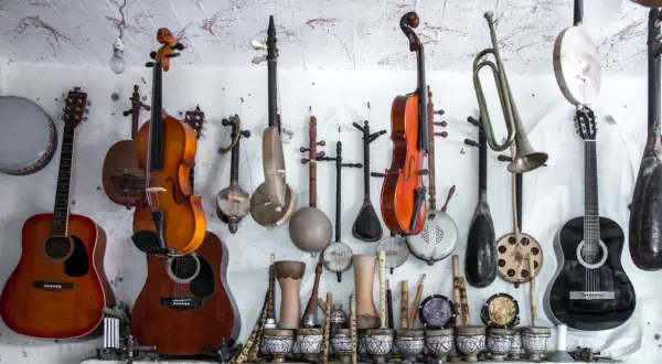 Różnorodne instrumenty strunowe, w tym gitary, skrzypce i tradycyjne lutnie, wiszą na ścianie obok bębnów i fletów.