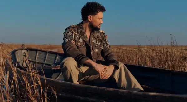 Calum Scott w łodzi, w kurtce ozdobionej broszkami, jakby śpiewał "You Are The Reason".
