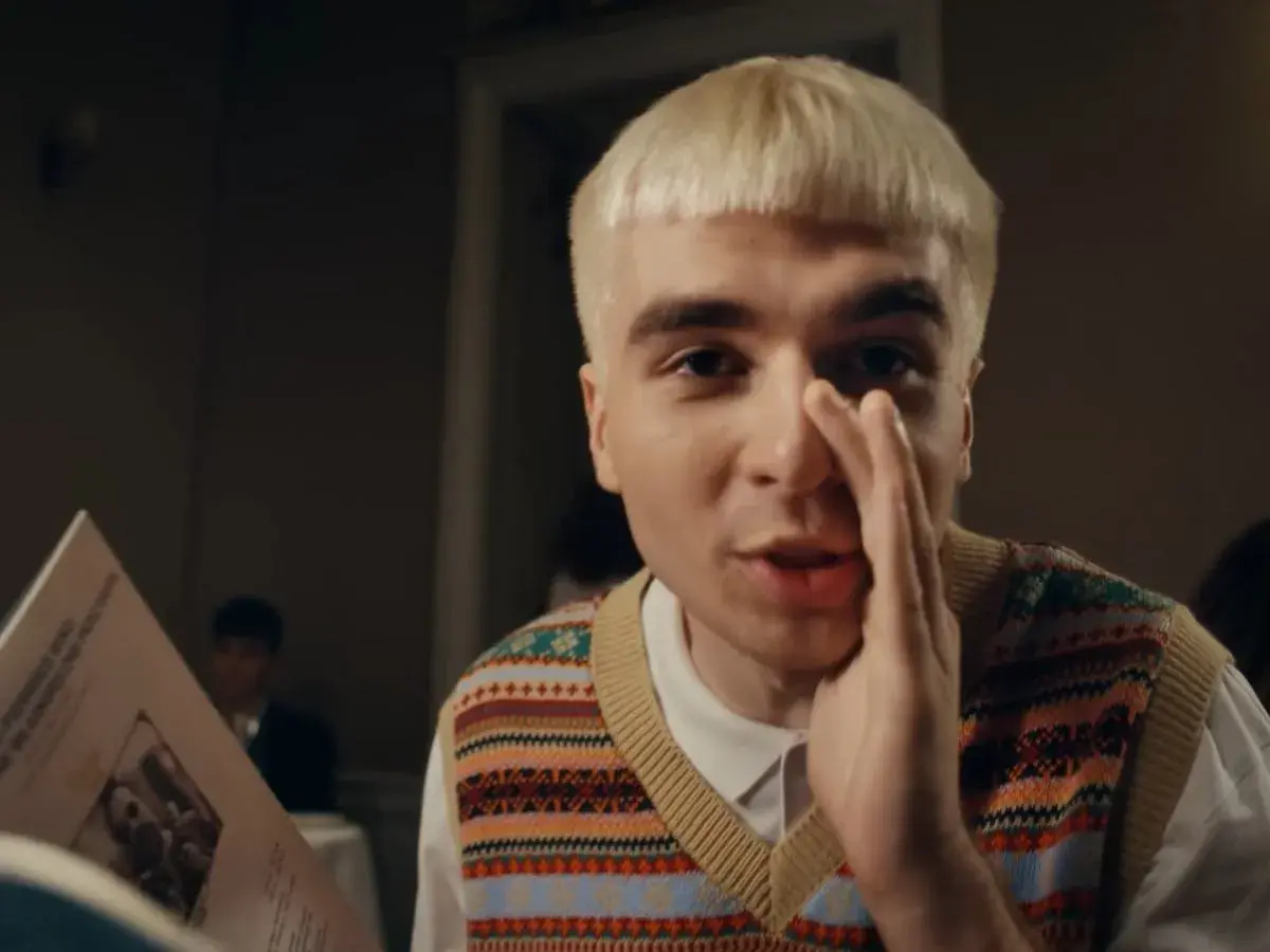 Młody mężczyzna z blond włosami, w kamizelce w kolorowe wzory, trzyma w ręku kartkę z tekstem, jakby czytał papuga.