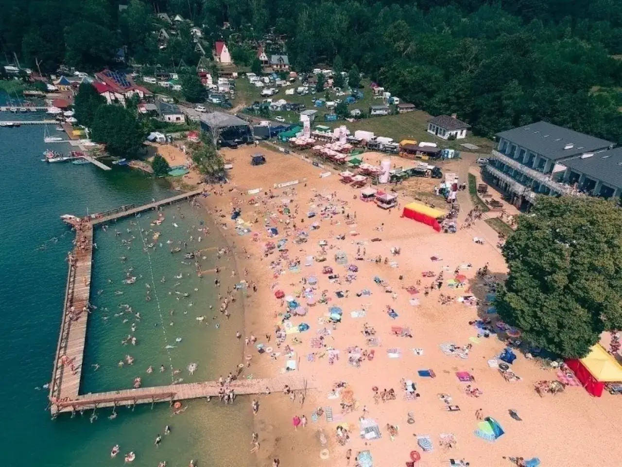 Tłum ludzi na plaży i w wodzie, z tamtej strony jeziora widać zabudowania i las.